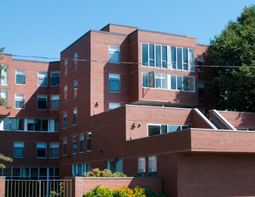 Brick building with multiple levels, windows on side of building, greenery in the foreground, blue skies in the background.