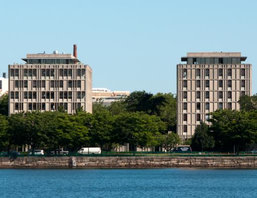 Two high rise buildings next to each other, greenery in front of the building, Charles river in the foreground, and blue skies in the background