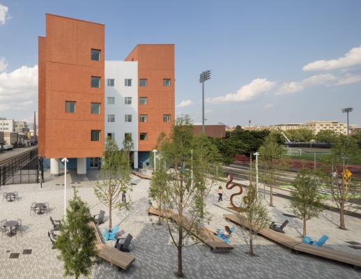 Orange and white building to the left, with outdoor lounge area with concrete ground and benches and chairs, greenery throughout the ground, blue skies in the background.